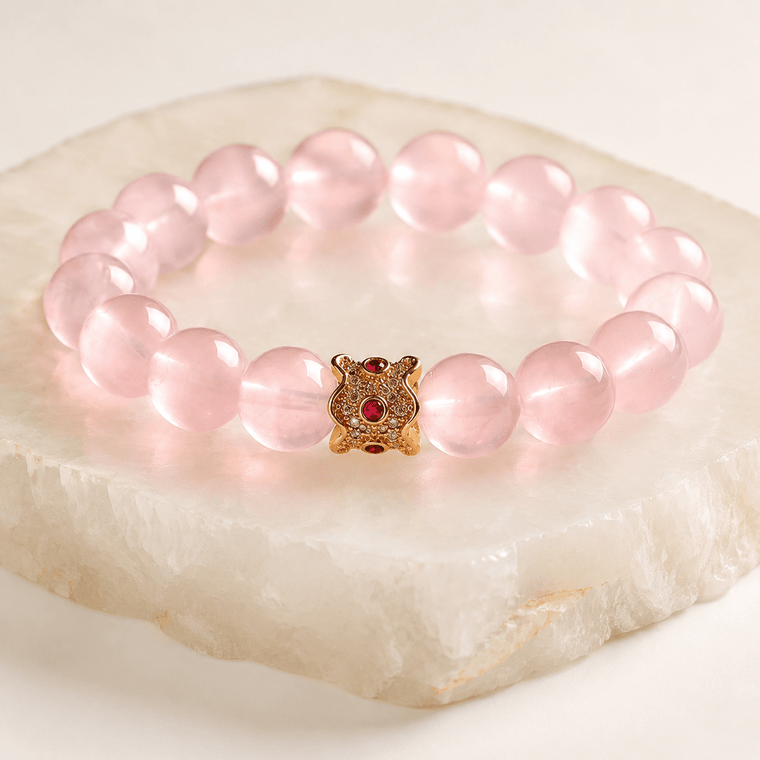 My Heart Is Safe Rose Quartz bracelet displayed on a pale stone slab with gold-tone ruby-red centerpiece